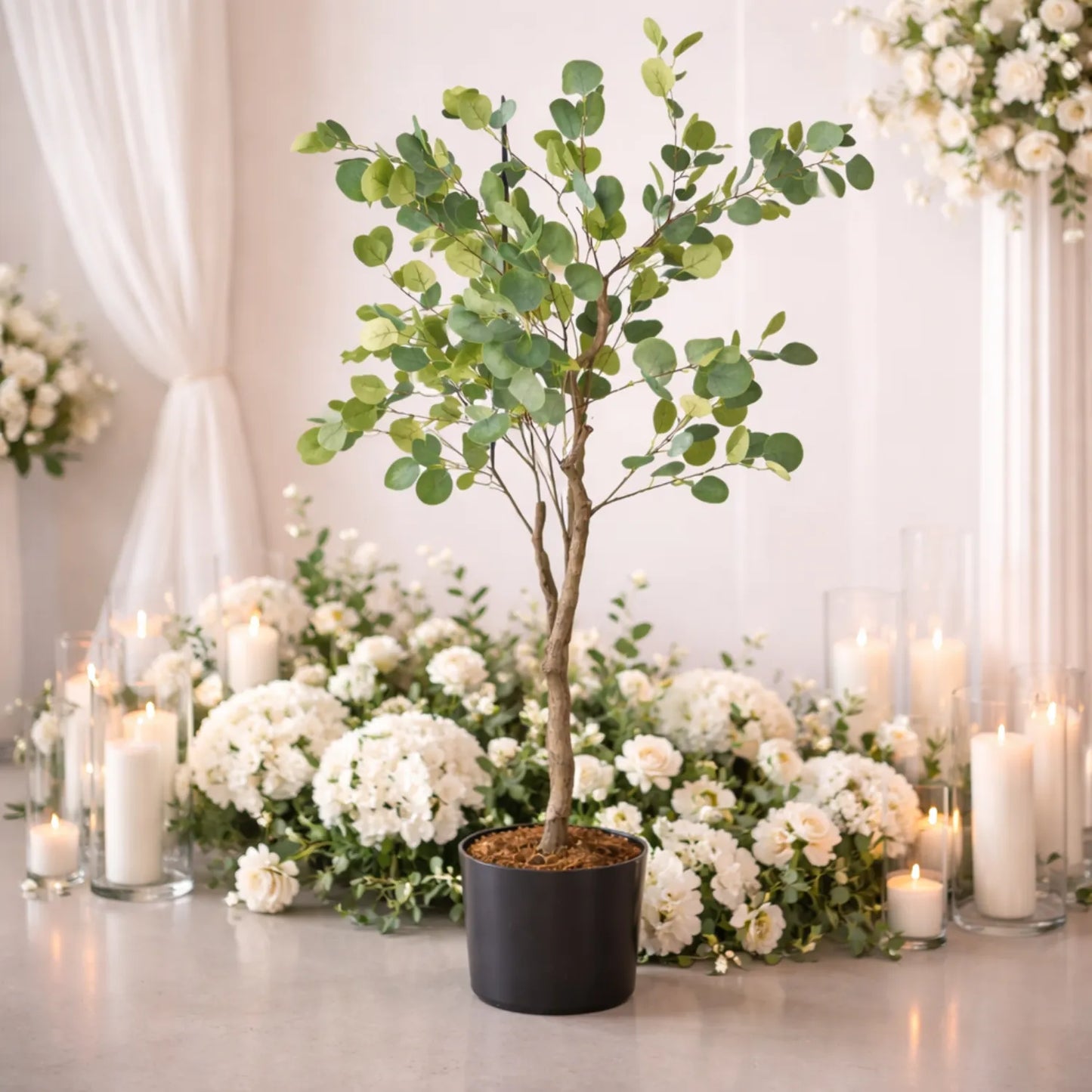Decorative setup with a potted tree and white flowers on a table, surrounded by candles.