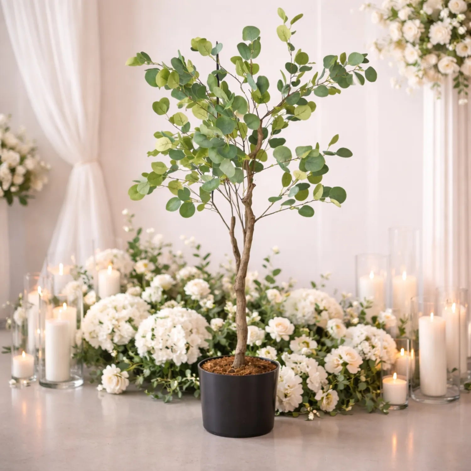 Decorative setup with a potted tree and white flowers on a table, surrounded by candles.