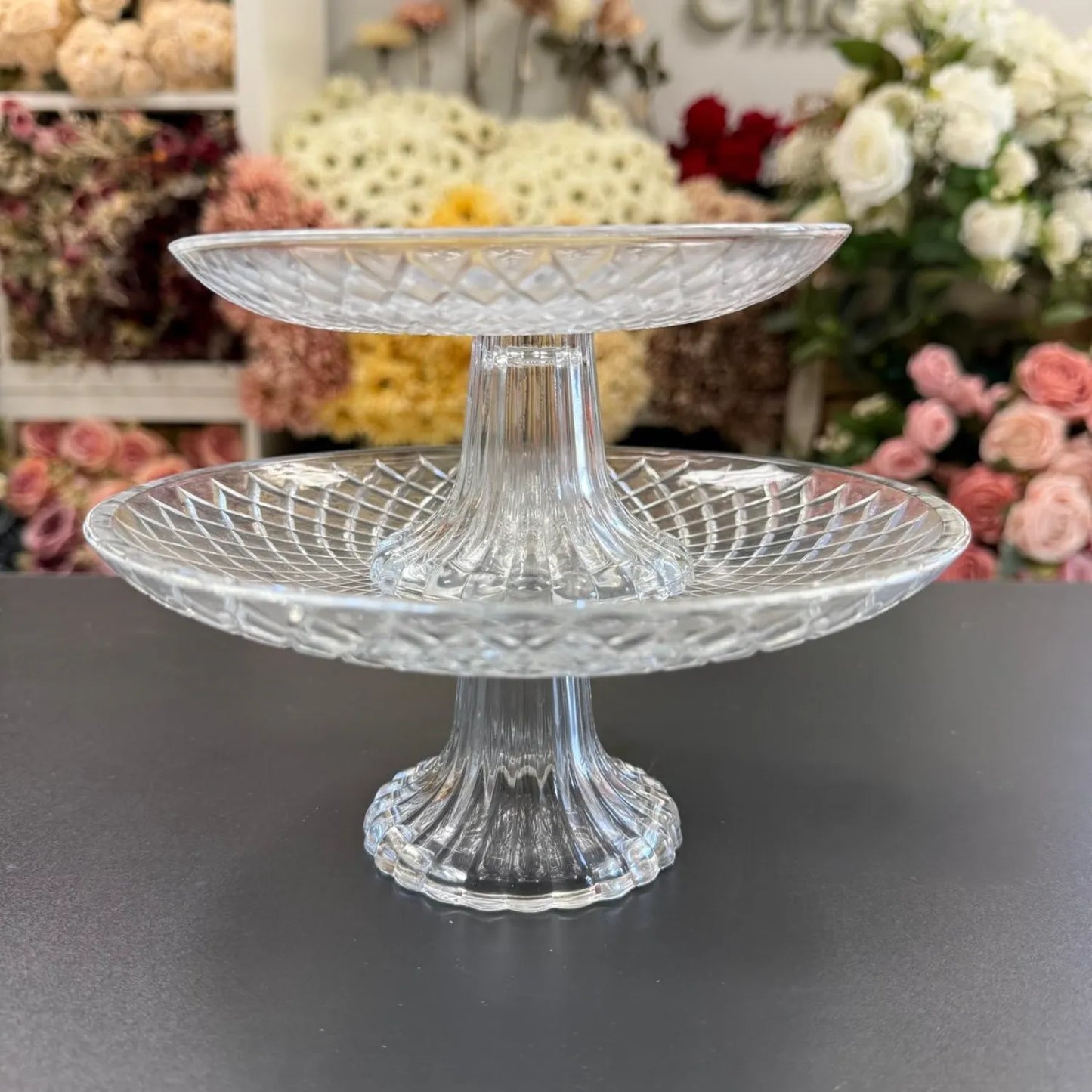 Clear glass two-tiered stand on a dark surface with floral decorations in the background