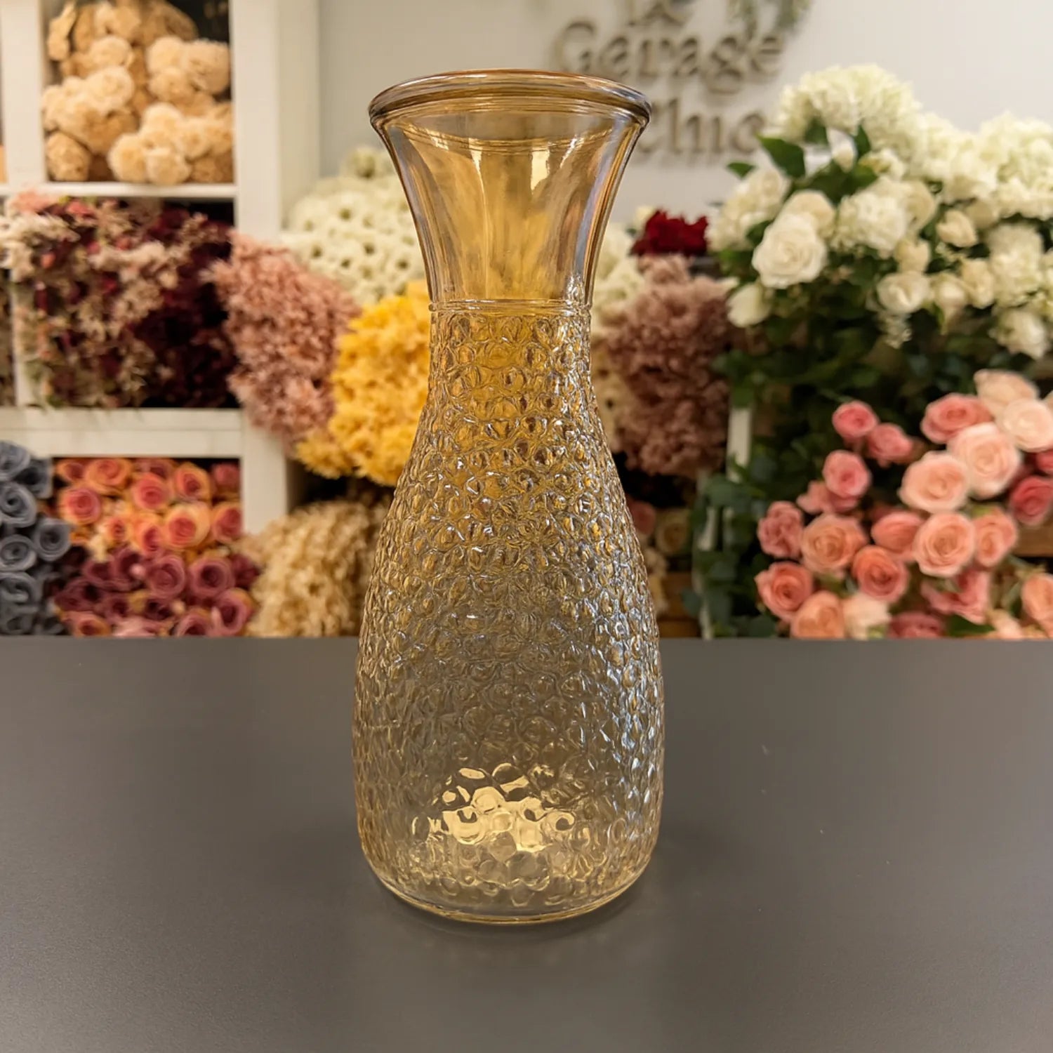 Decorative glass vase with textured design on a gray surface, flowers in the background