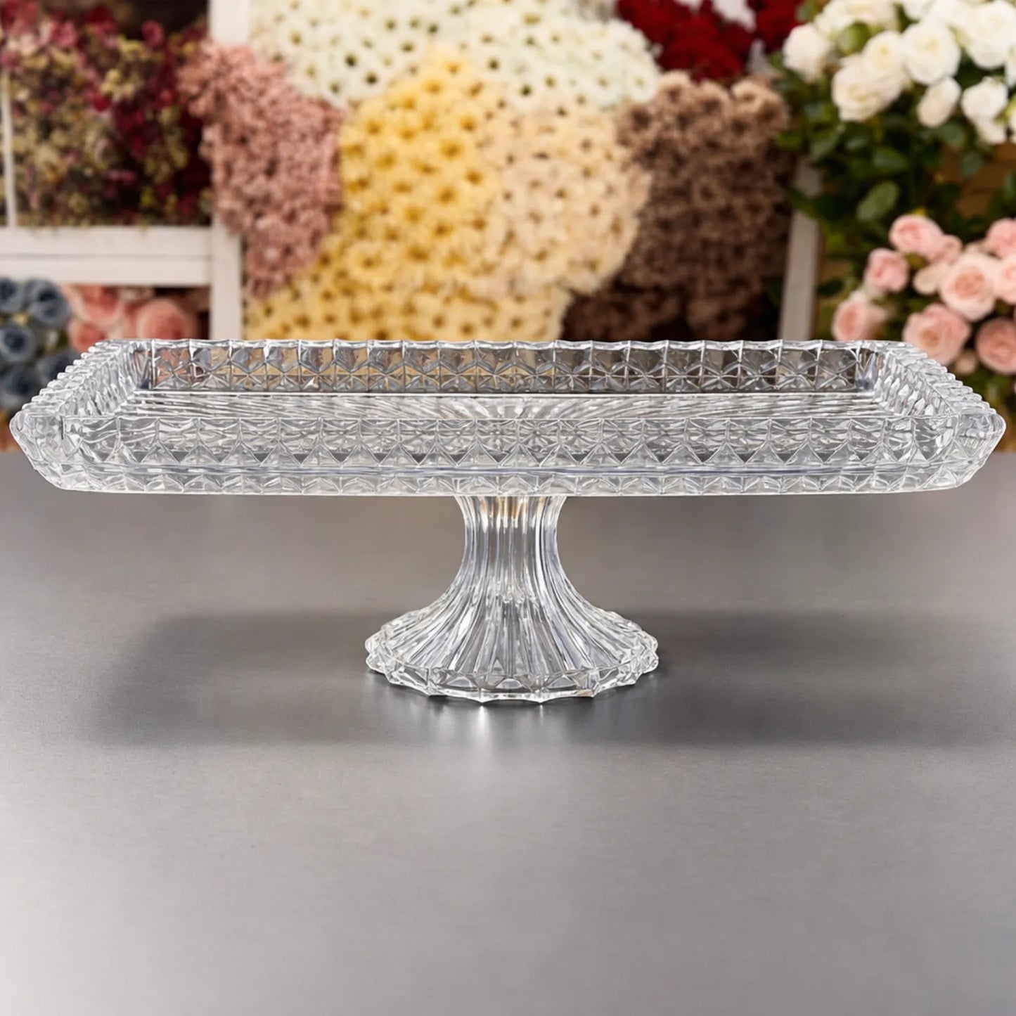 Clear glass cake stand on a gray surface with floral decorations in the background