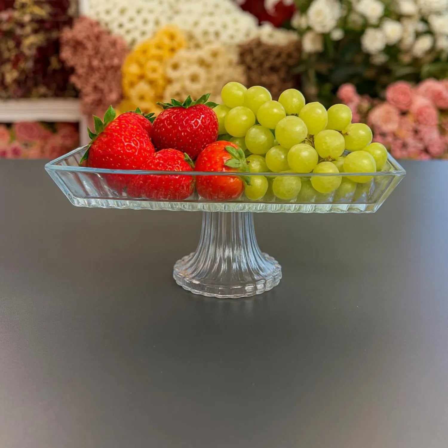Clear glass fruit bowl with strawberries and grapes on a gray surface, with blurred floral background