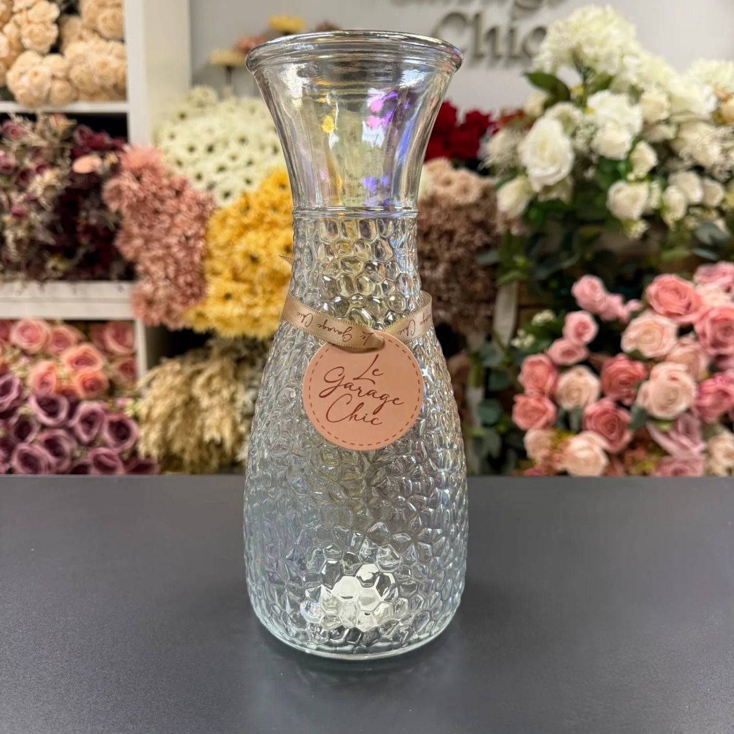 Clear textured vase with a label on a dark surface, flowers in the background