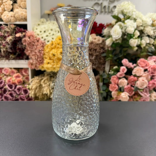 Clear textured vase with a label on a dark surface, flowers in the background