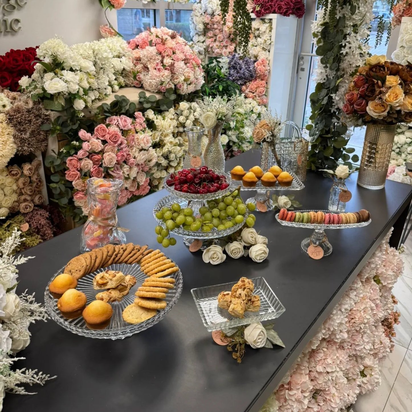 Table with assorted pastries and fruits surrounded by floral arrangements in a decorated setting.